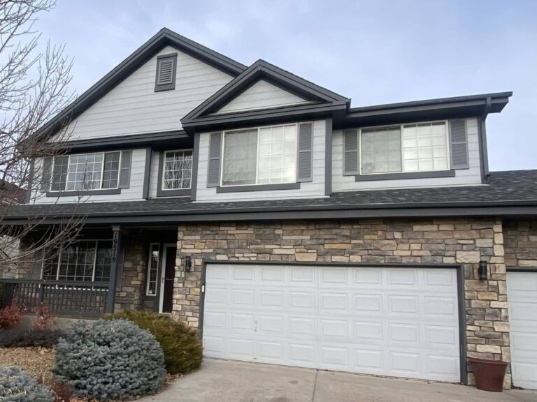 Two-story suburban house with stone and gray siding exterior, white-framed windows, and a three-car garage. There are shrubs and a leafless tree in the front yard.