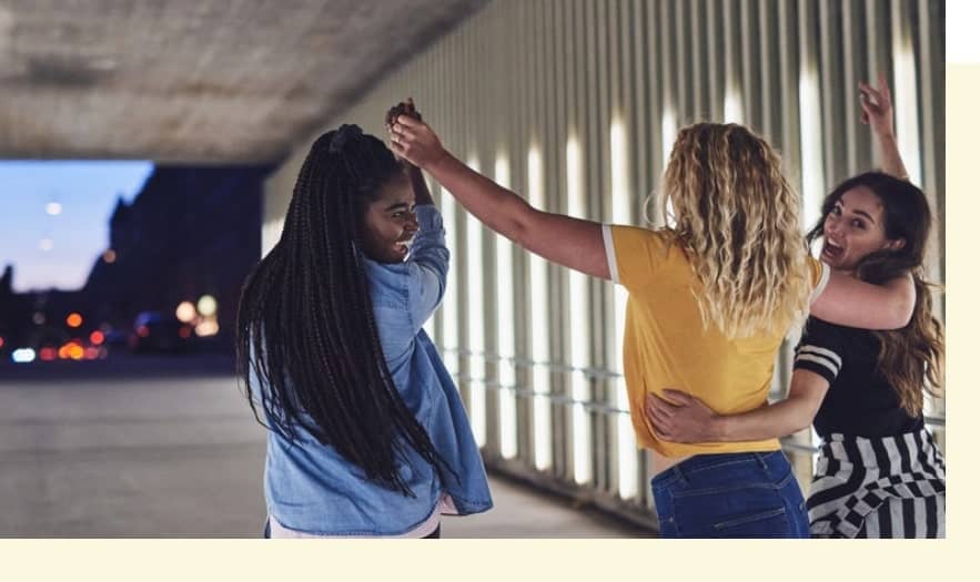 Three women in casual clothing dance joyfully under a well-lit bridge or walkway in the evening.