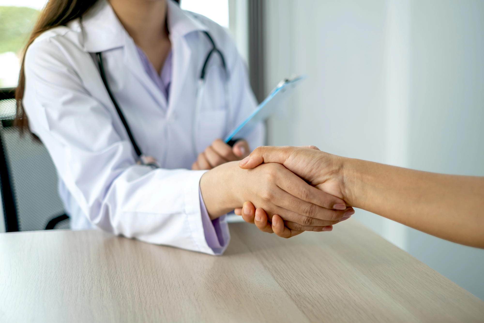 A doctor wearing a white coat and stethoscope shakes hands with a patient across a desk, holding a clipboard in the other hand.