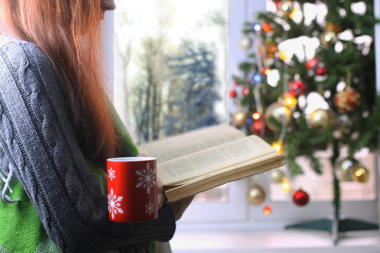 A person with long hair reads a book next to a window. They hold a red mug with snowflake designs and a decorated Christmas tree is in the background.