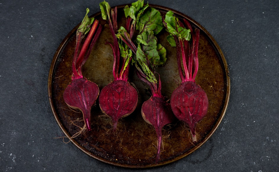 Halved beets with greens on a round metal tray placed on a dark surface.
