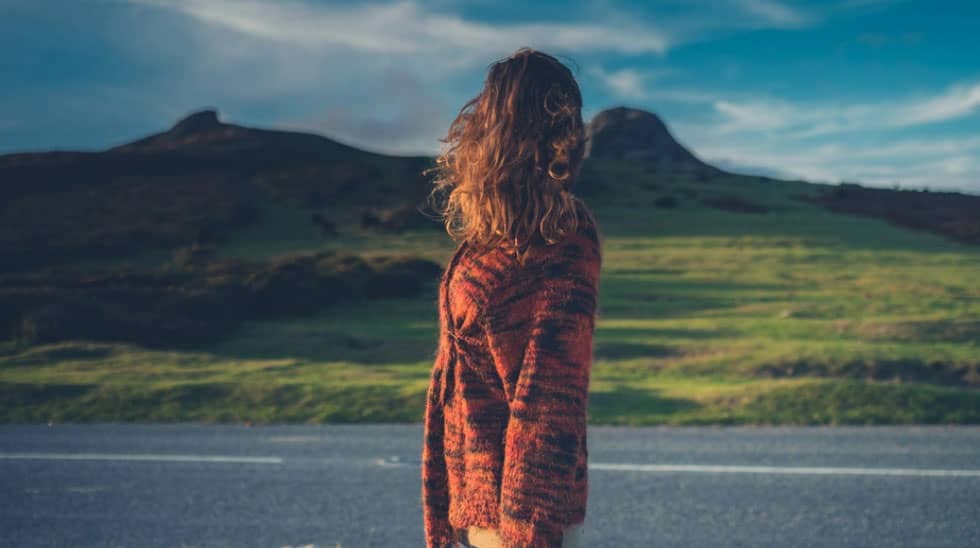 Person with long hair in a red sweater standing near a road, looking at green hills and distant peaks under a blue sky.