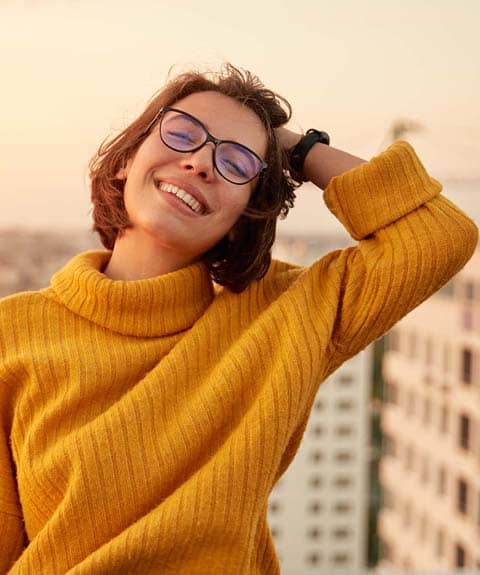 A person with short hair and glasses smiles while wearing a yellow sweater, standing outdoors with buildings in the background.