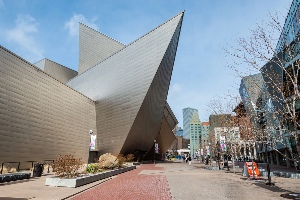 A modern, angular building is shown with sharp, pointed edges, flanked by an open plaza. The sidewalk and street are visible, with sparse vegetation and leafless trees lining the path.