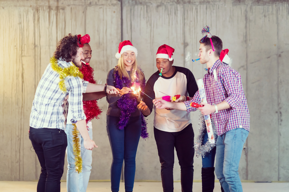 Five people wearing festive hats and tinsel, holding sparklers and party items, celebrating indoors.