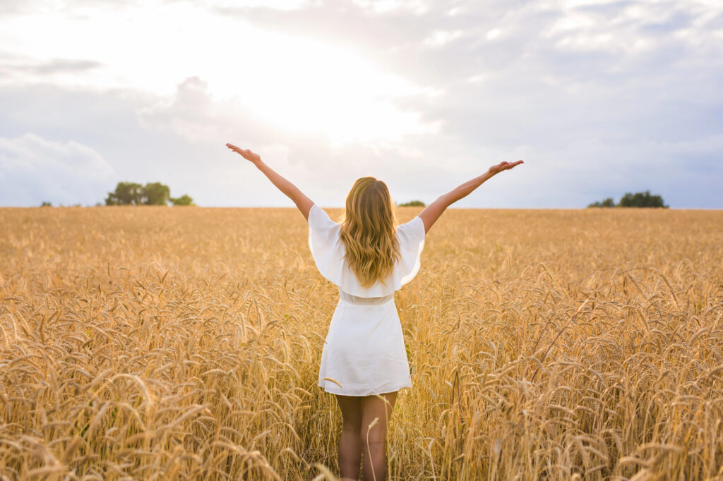 A woman in a white dress stands in a wheat field with arms raised, facing the sun and cloudy sky.