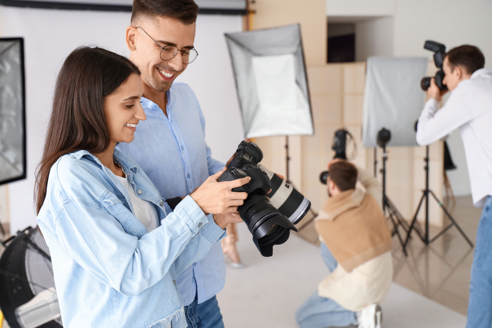 A man and woman look at a camera screen in a photography studio, while two other people work with cameras in the background.