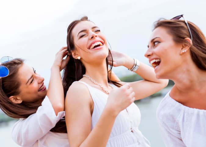 Three women wearing white tops laugh and enjoy themselves outdoors with a blurred natural background.