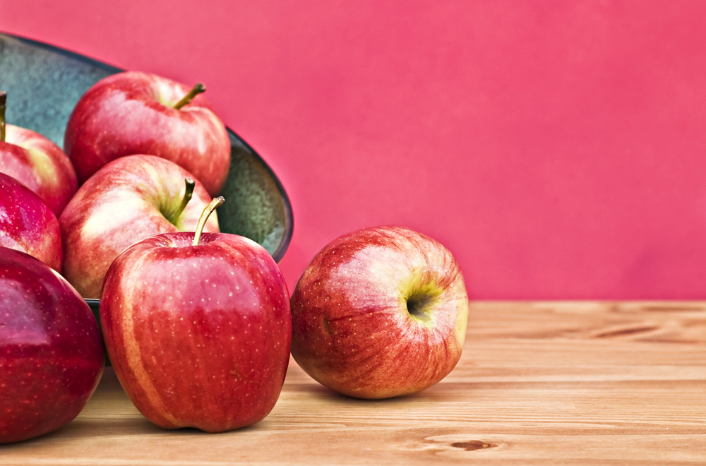 A bowl of red apples on a wooden surface, with two apples outside the bowl 