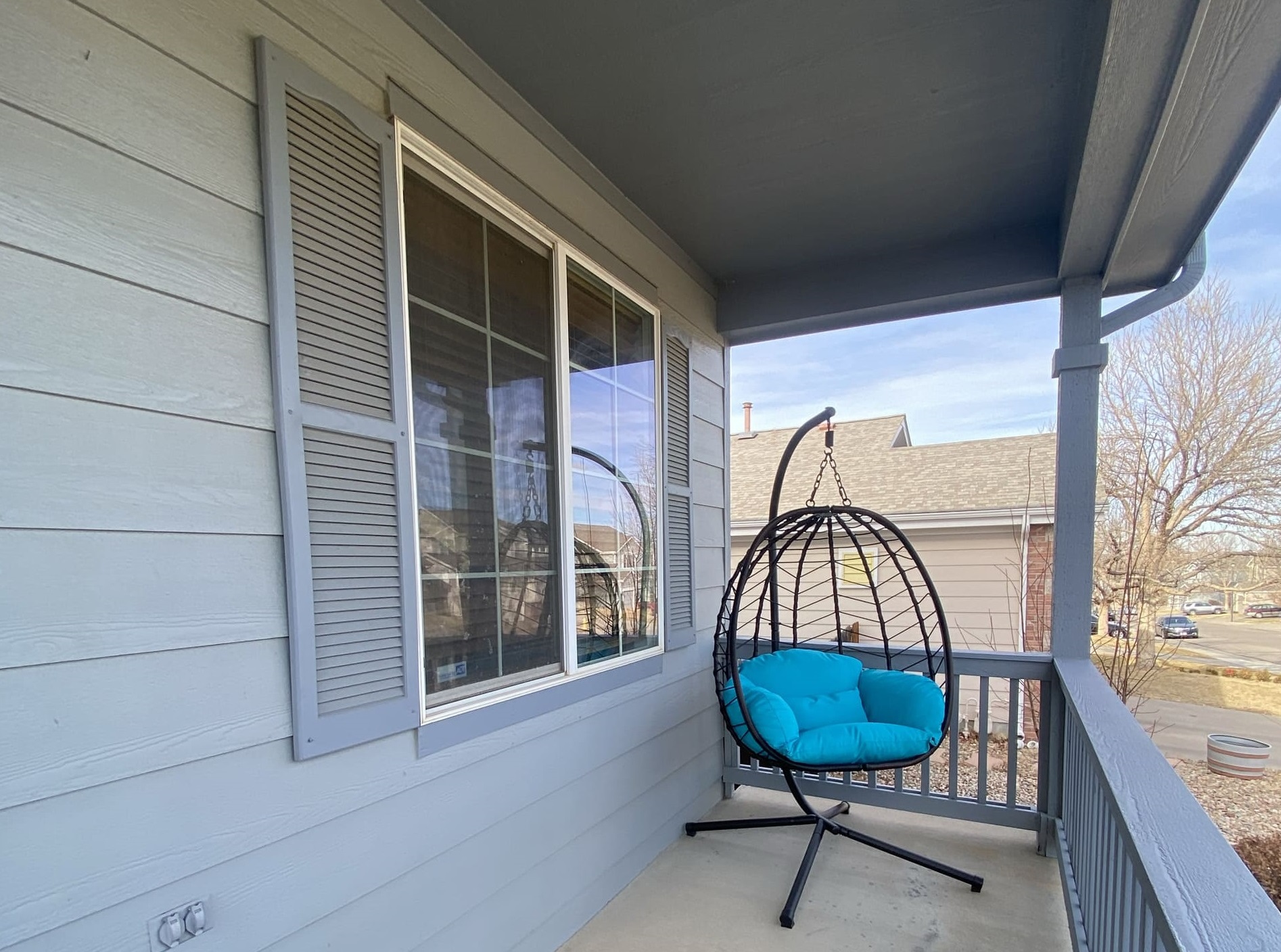 A porch with a gray exterior, featuring a hanging egg chair with turquoise cushions. Nearby are a window with shutters and a view of houses and trees.