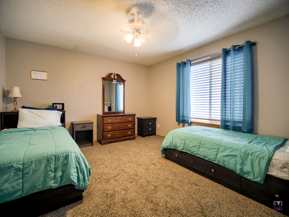 A bedroom with two twin beds, each with teal bedding, a dresser with a mirror between them, and a window with blue curtains. The room has a ceiling fan and a carpeted floor.