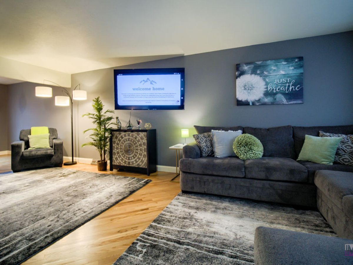 Modern living room with gray sofas, a patterned rug, wall-mounted TV, floor lamp, potted plant, and decorative wall art reading "Just breathe.
