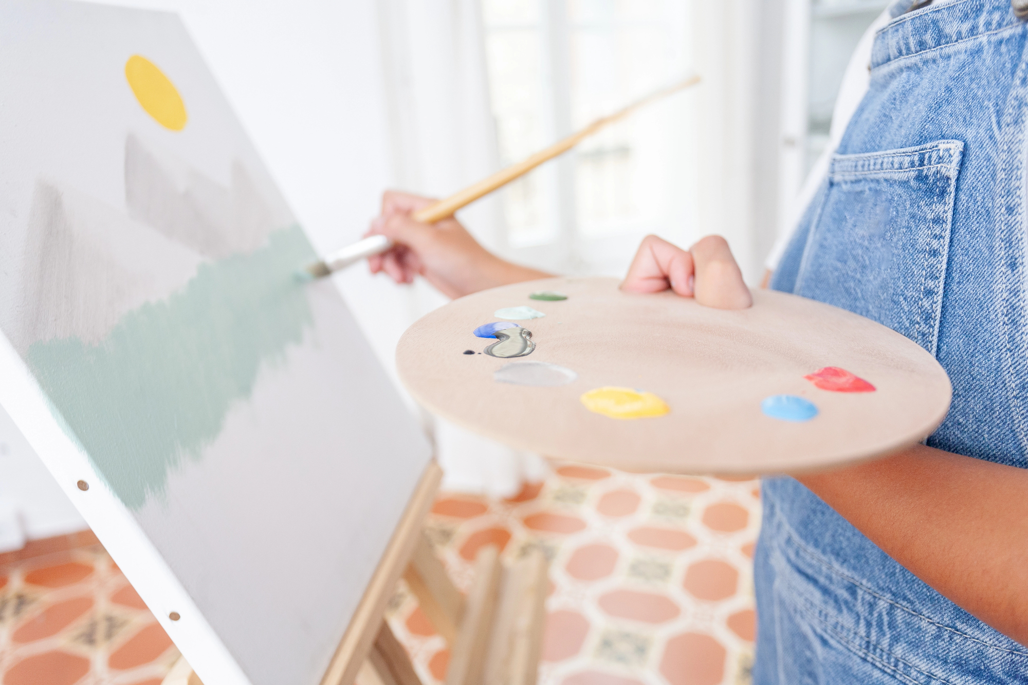 Person in denim holding a paint palette and painting a landscape with mountains, sun, and water on a canvas using a paintbrush.