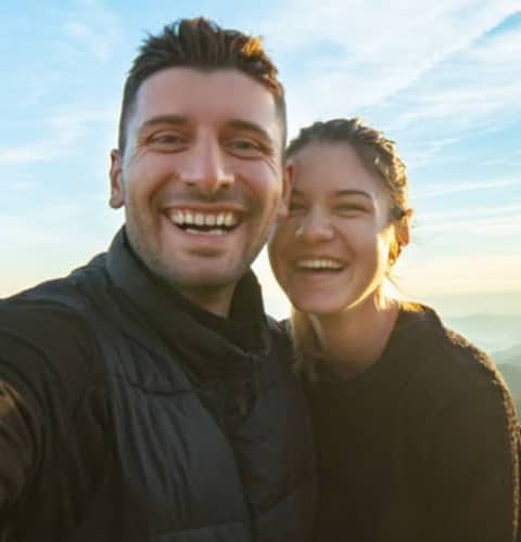 A man and a woman smiling at the camera, standing closely together with a clear sky in the background.