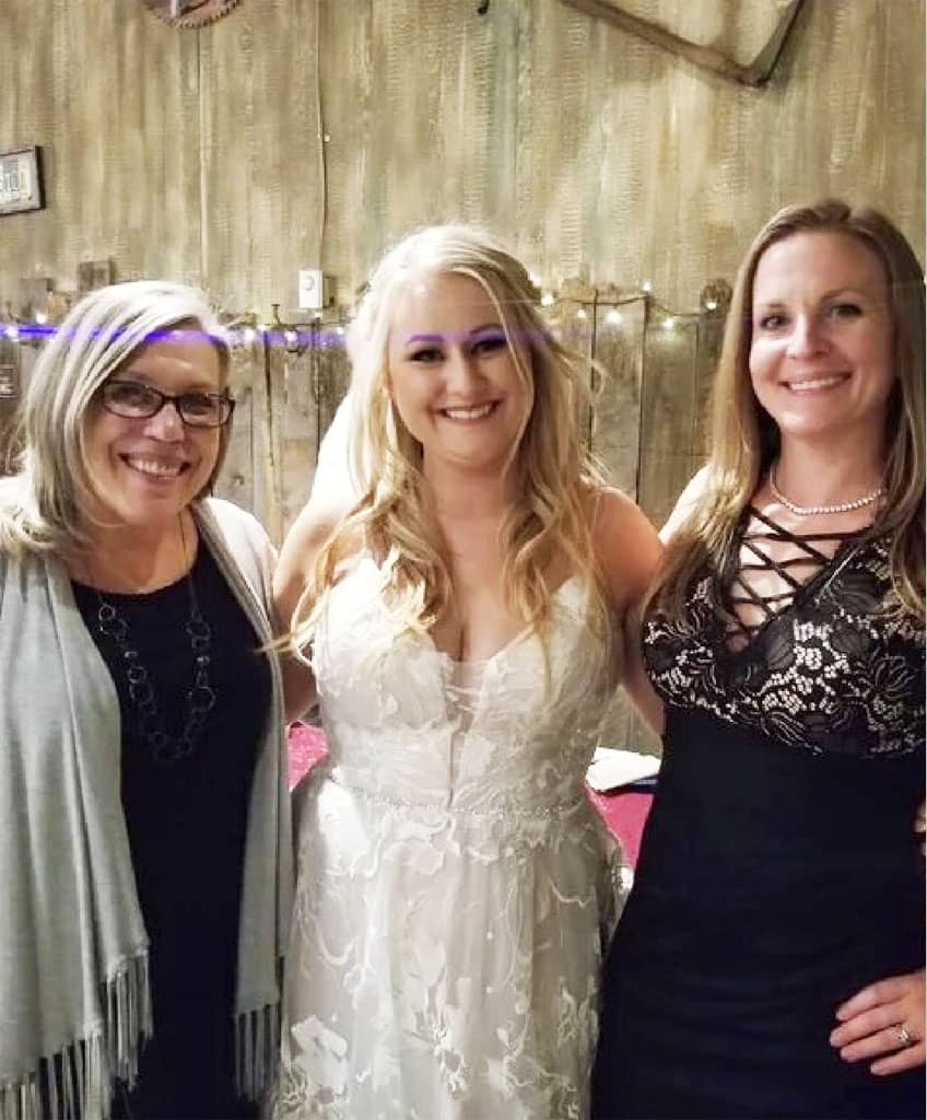 Three women pose together: one in a grey cardigan, one in a white lace wedding gown, and one in a black dress with a crisscross neckline. They stand in front of a rustic wooden backdrop.