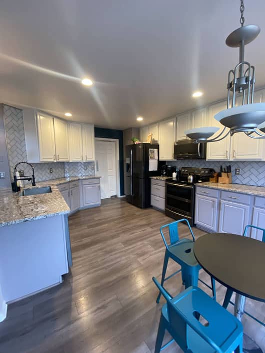 Modern kitchen with white cabinets, black appliances, grey countertops, and wooden flooring. Blue chairs around a small round dining table under a hanging light fixture.