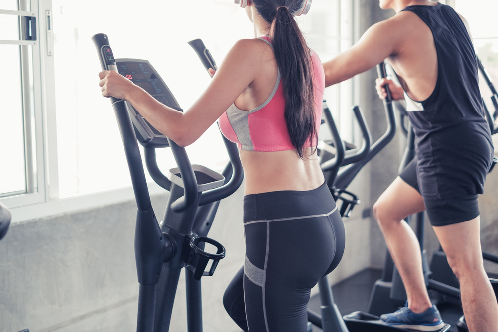 Two people, one in a pink sports bra and another in a black tank top, use elliptical machines in a gym