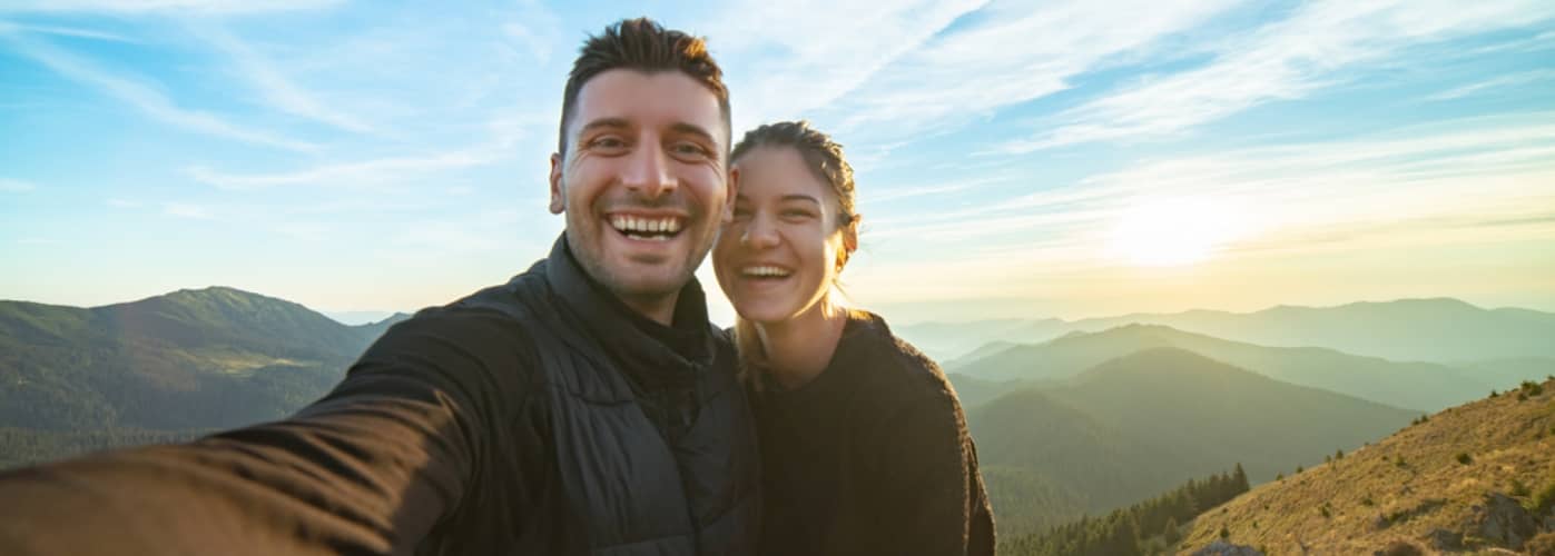 A smiling couple takes a selfie on a mountain peak with a scenic view of hills and a sunset in the background.