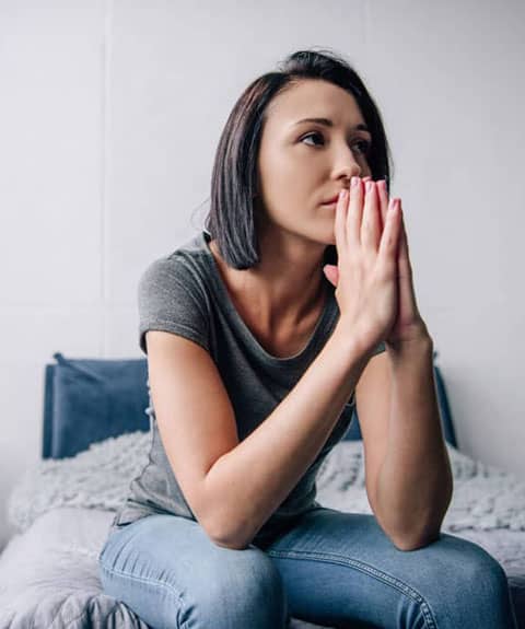 A woman with short dark hair and a grey t-shirt sits on a bed with her hands pressed together near her face, appearing thoughtful or contemplative.