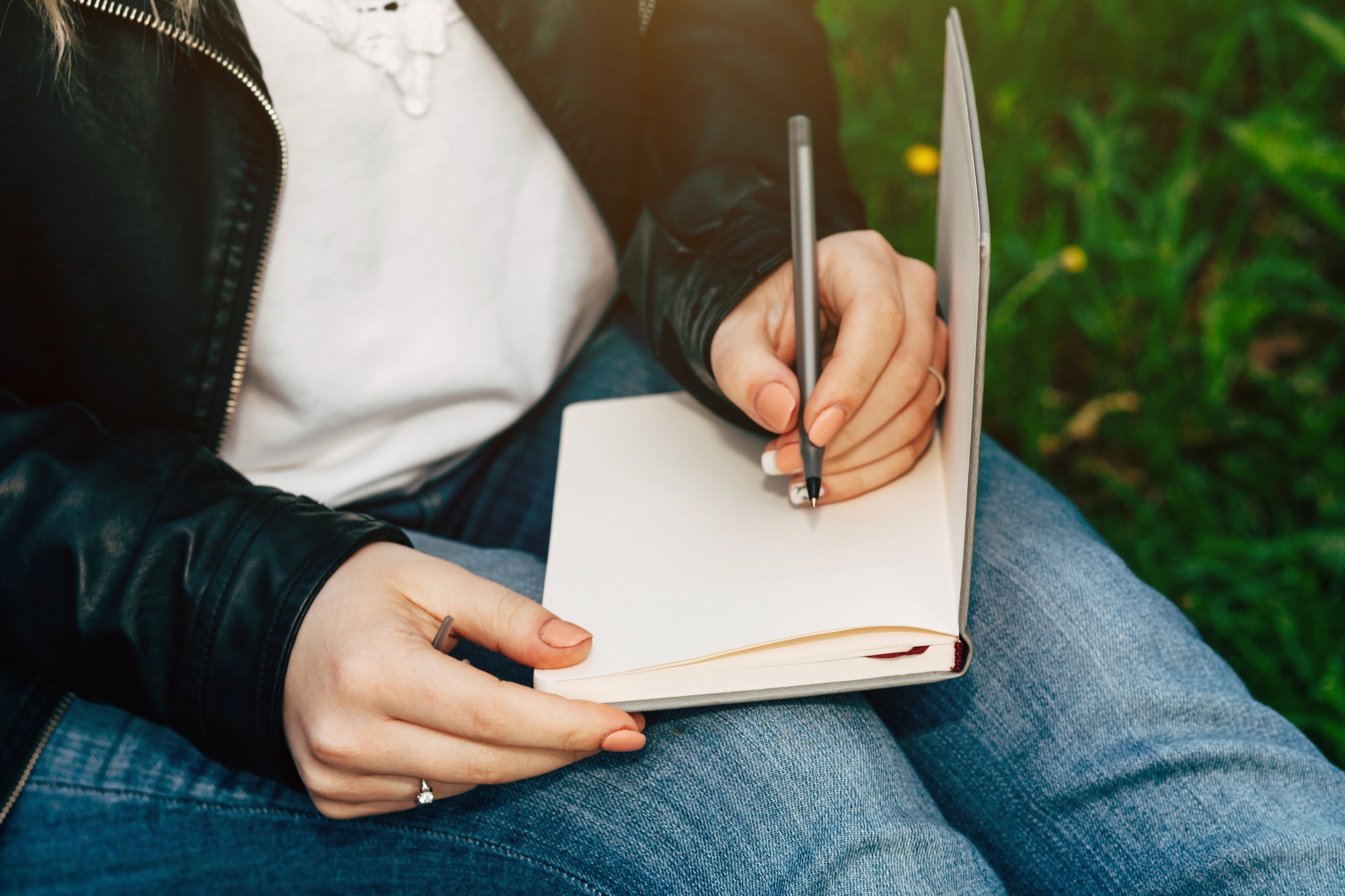 A person sits outdoors on grass, wearing jeans and a black jacket, writing in a blank notebook with a pen.