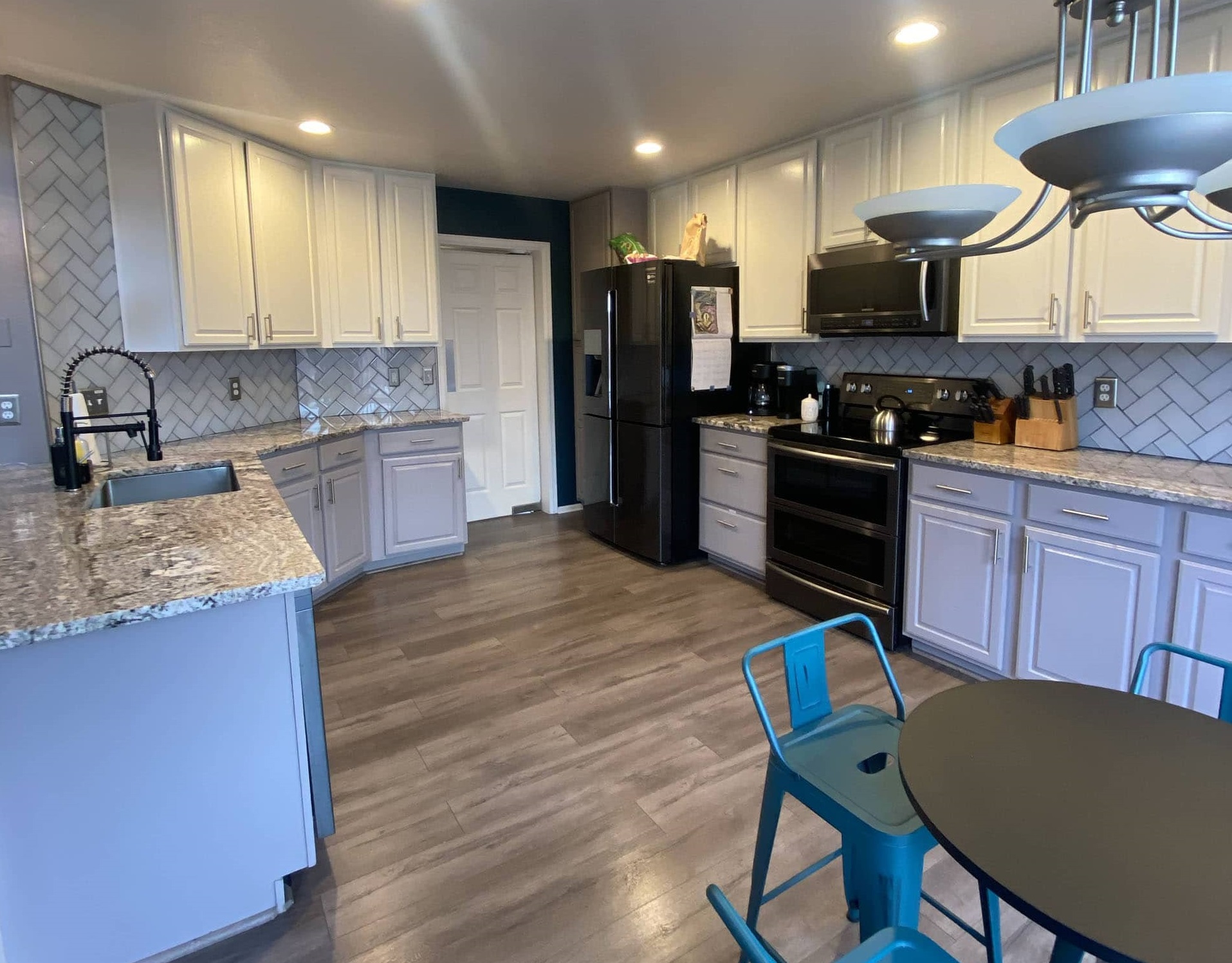 Modern kitchen with granite countertops, black appliances, gray cabinets, and wood flooring. Blue metal chairs are around a black round table. 