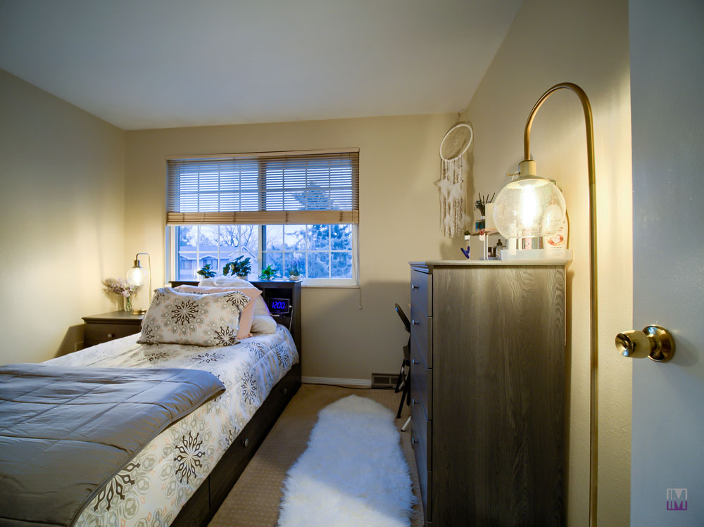 A cozy bedroom with a bed, dresser, and window. The room has a soft rug, lamp on the right, and a dreamcatcher on the wall. Neutral-colored walls and bedding create a calm atmosphere.