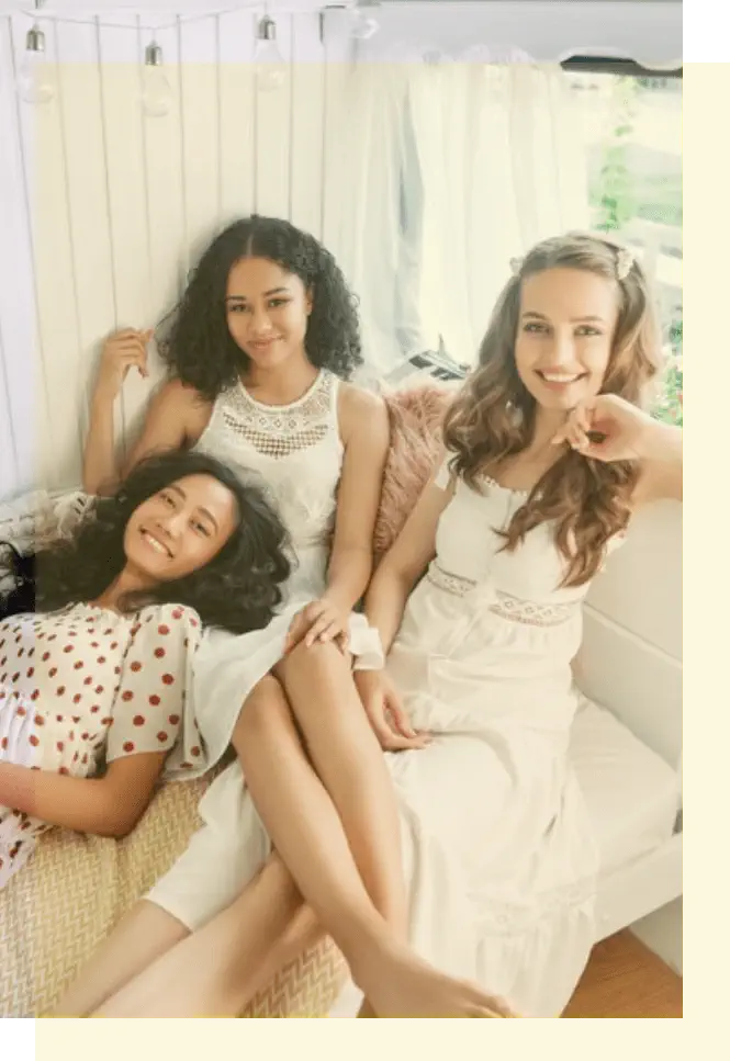 Three women sit and smile on a white couch by a window. Two wear white dresses, and one wears a white dress with red polka dots. The setting is bright and airy.