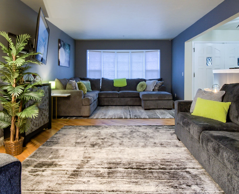 A modern living room featuring grey sofas with green accents, a large window with blinds, and a potted plant. A rug covers the wooden floor, and artworks adorn the walls.
