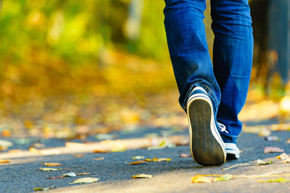 Person wearing blue jeans and sneakers walks on a path covered with fallen leaves in an outdoor setting.