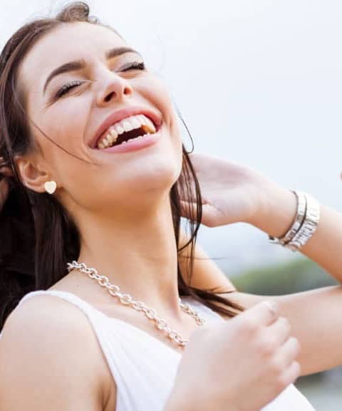 A woman with dark hair and heart-shaped earrings is joyfully laughing, wearing a white top and silver necklace, with one arm raised and the other brushing her hair back.