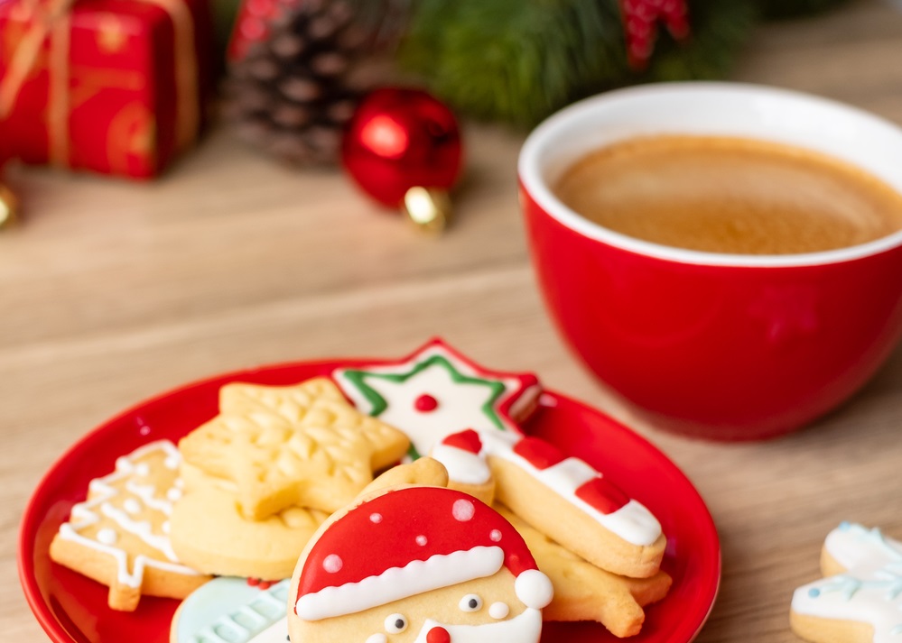 A red plate with Christmas-themed cookies, including a Santa face, next to a red cup of coffee on a wooden table. Holiday decorations are blurred in the background.