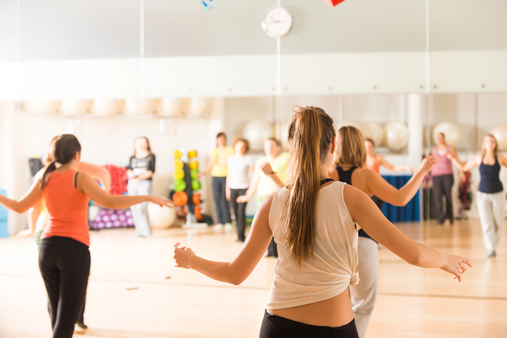 People participating in a group dance or fitness class in a brightly lit room with mirrors and exercise equipment in the background.