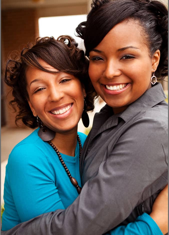 Two women smiling and embracing each other, one wearing a blue top with a necklace and the other in a gray top with earrings. They are in an outdoor setting with a blurred background.