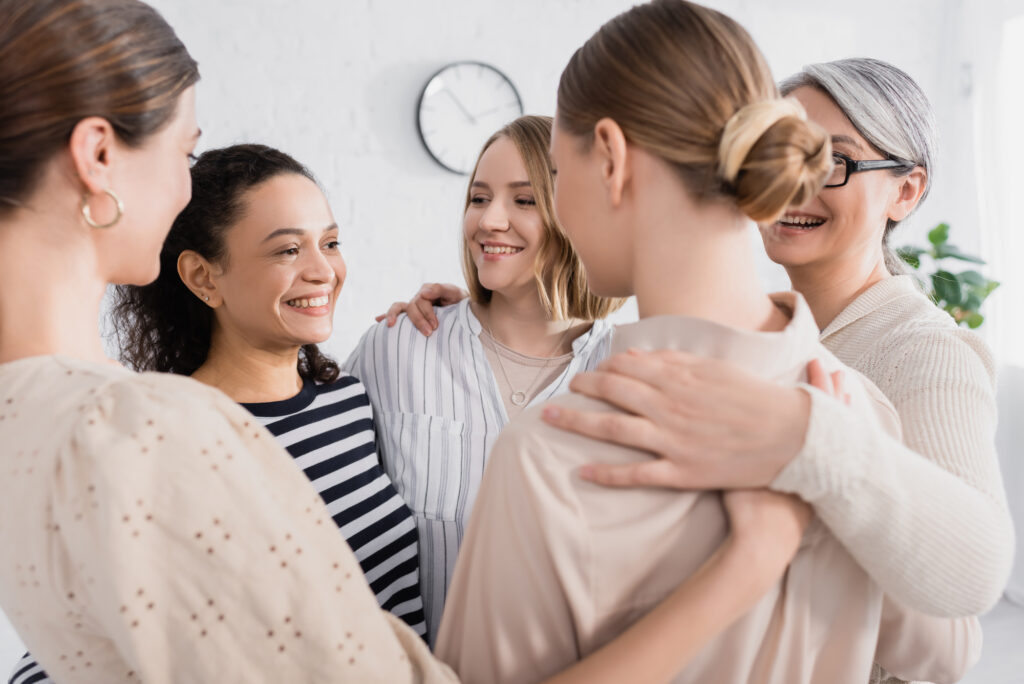 Five women stand in a close group, smiling and talking with their arms around each other in a bright room.