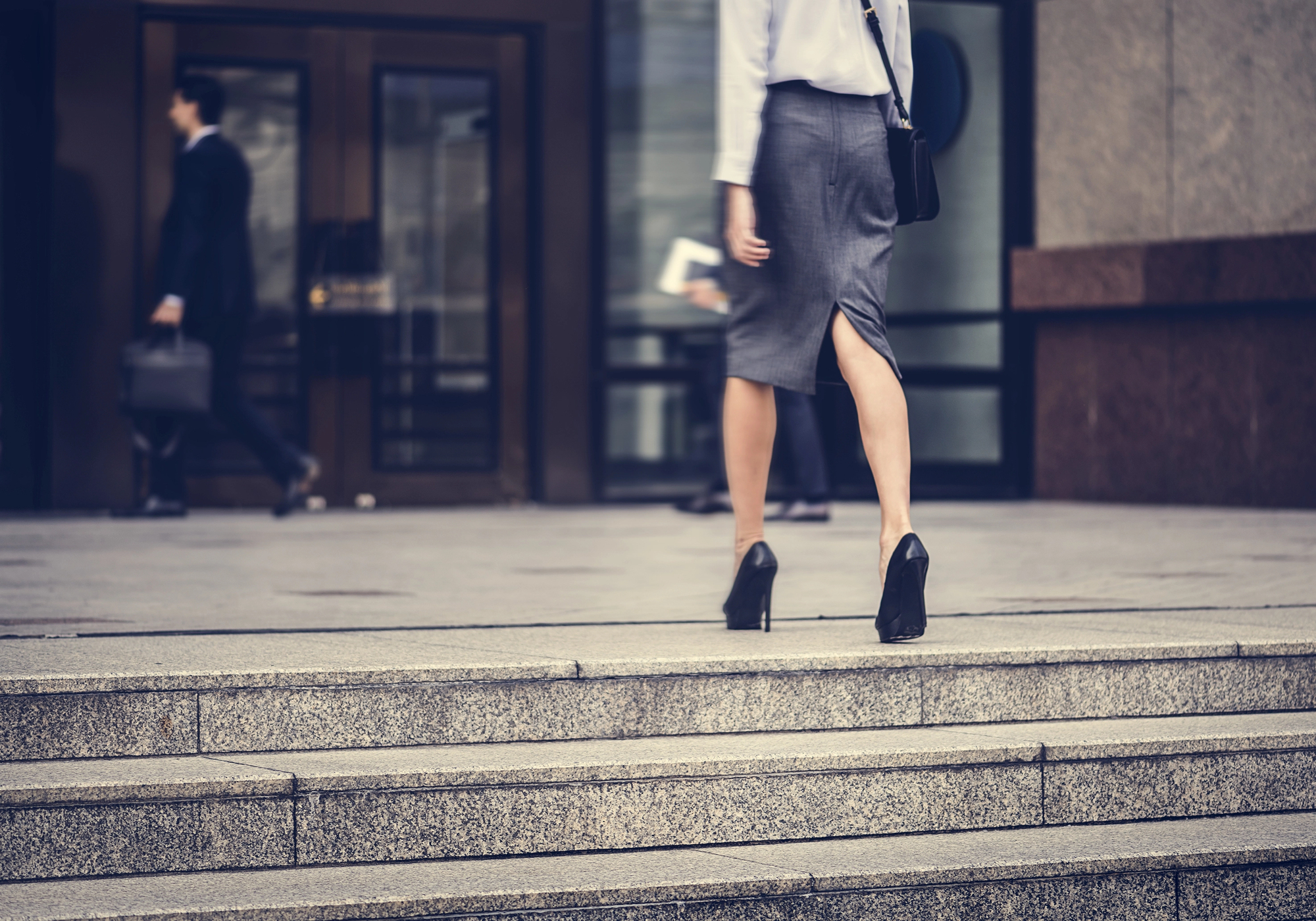 A person in a gray pencil skirt and black heels walks up concrete steps toward a glass building entrance.
