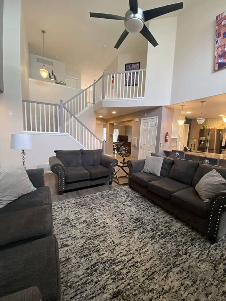 A living room with gray sofas, a patterned rug, and a visible staircase leading to an upper level. The room features a ceiling fan and pendant lights, with a kitchen area in the background.