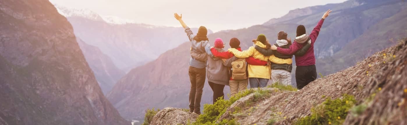 A group of seven people wearing jackets stands on a rocky ledge in front of a mountainous landscape, with their arms raised or around each other.