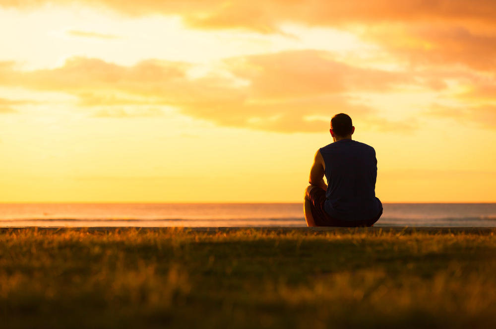 A person sits on the grass facing the ocean, watching the sunset with an expansive sky in the background.
