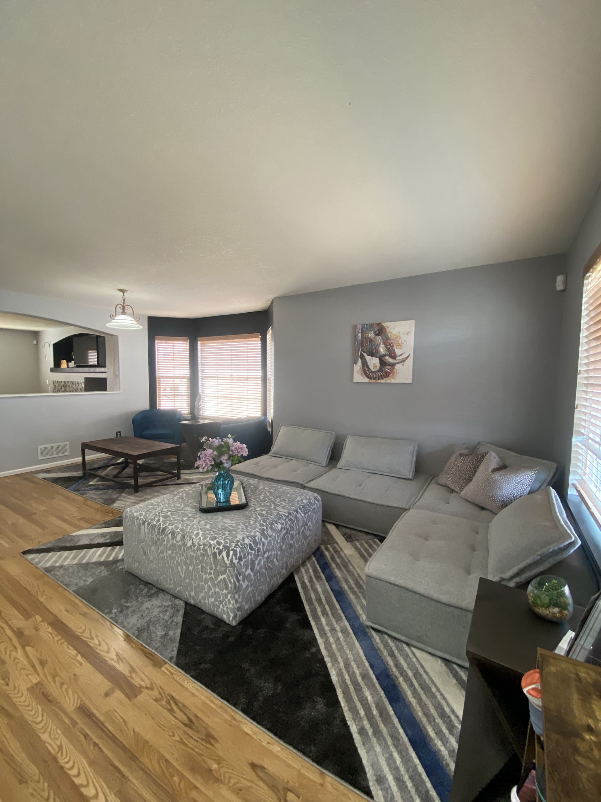 A modern living room with a gray sectional sofa, a geometric-patterned ottoman, a dark coffee table, and a wall-mounted decorative picture. Wooden floors and light gray walls are visible.