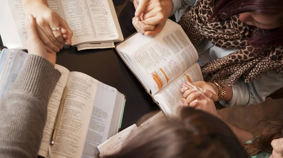 A group of people sitting around a table holding hands, with open books in front of them.