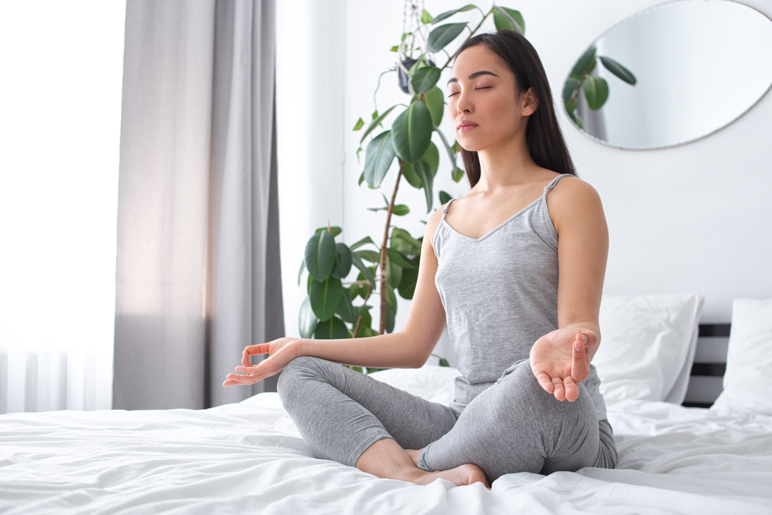 A person sits cross-legged on a bed in a meditative pose, eyes closed, dressed in gray loungewear, with a potted plant and a mirror in the background.