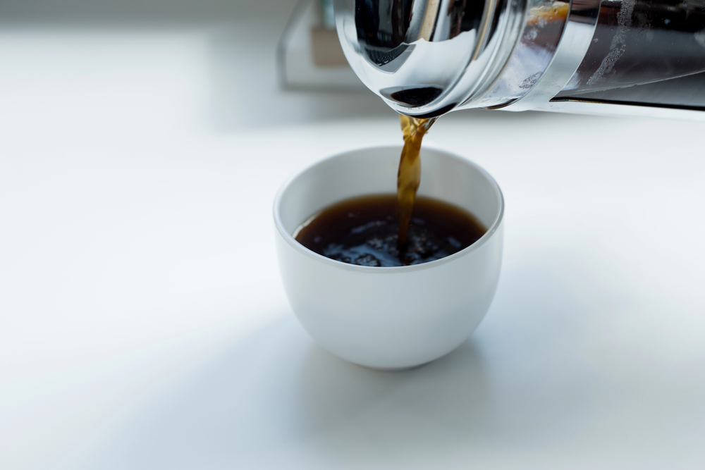 Coffee being poured from a glass pot into a white ceramic cup on a white surface.