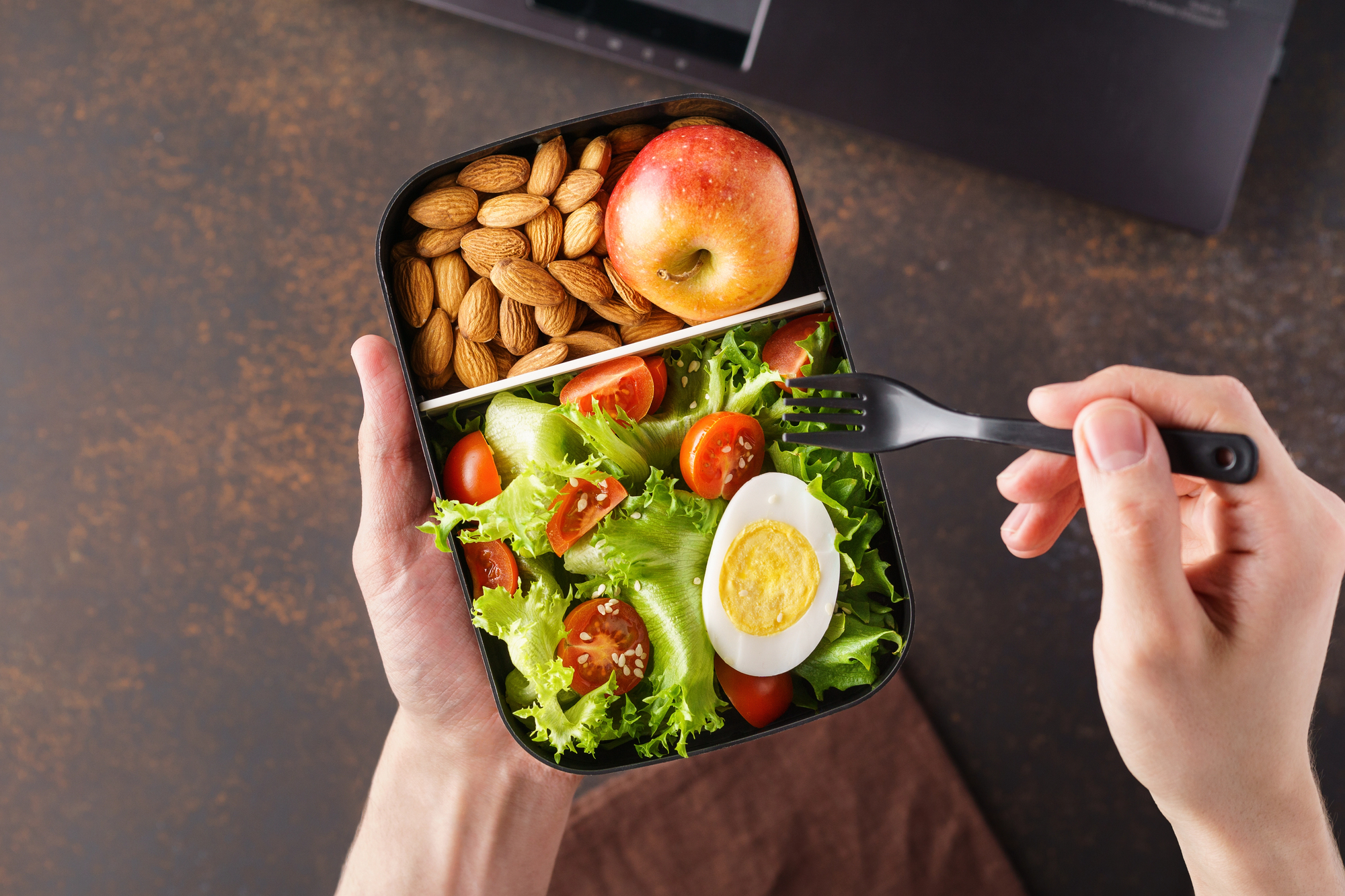 A person holds a lunchbox containing almonds, an apple, and a salad with lettuce, cherry tomatoes, and a halved boiled egg, while using a black fork.