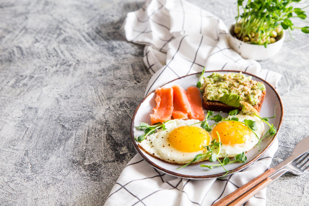 A plate with two sunny-side-up eggs, sliced smoked salmon, and avocado toast, served on a white cloth with a fork and knife beside it.