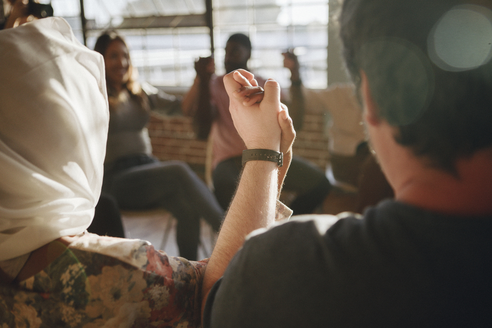 A group of people sit in a circle holding hands, during support group discussion