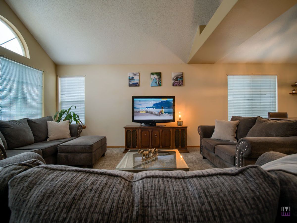 A living room with two gray sofas, a glass coffee table, a TV on a wooden stand, and three pictures on the wall above the TV. Light streams in from windows on both sides.