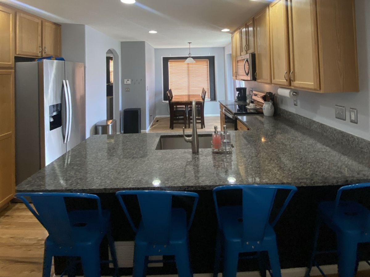 Modern kitchen with wood cabinets, stainless steel appliances, granite countertops, and four blue metal barstools at an island. Dining table visible in the background.