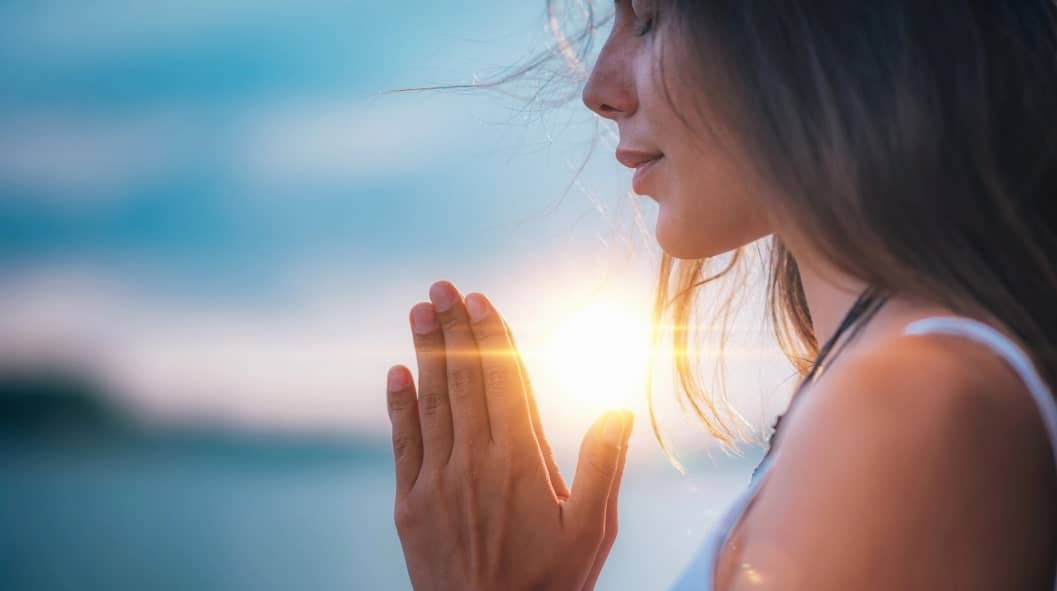 woman meditating with hands together and eyes closed with sunset view