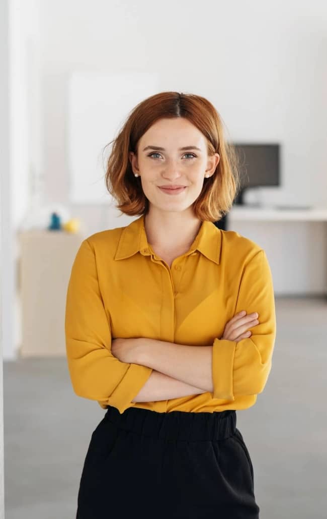 Happy relaxed confident young businesswoman standing with folded arms in a spacious office looking at the camera with a warm friendly smile