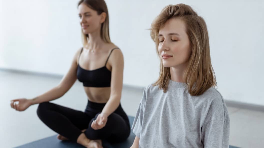 Two athletic attractive girls sit in lotus position on yoga mat indoors as part of benefits of sober living